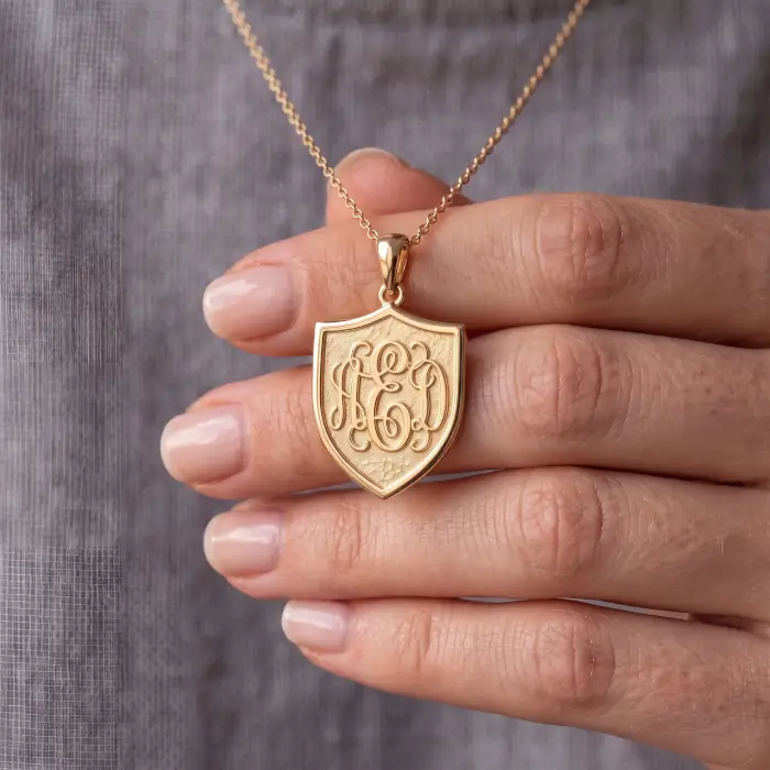 A hand holding a custom engraved gold shield necklace featuring an intricate script monogram. This shows the high-precision results achievable on fine jewelry using our clean, continuous-line vector files.