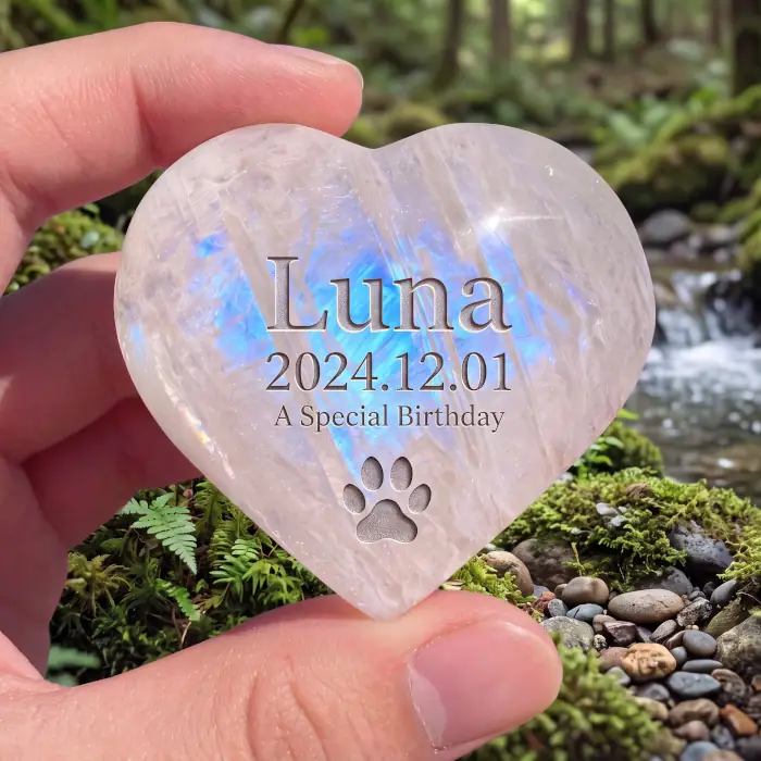 A hand holding a heart-shaped moonstone engraved with a pet's name, date, and a paw print. This showcases the precision achievable on crystal and stone using our custom vector text and icon files.